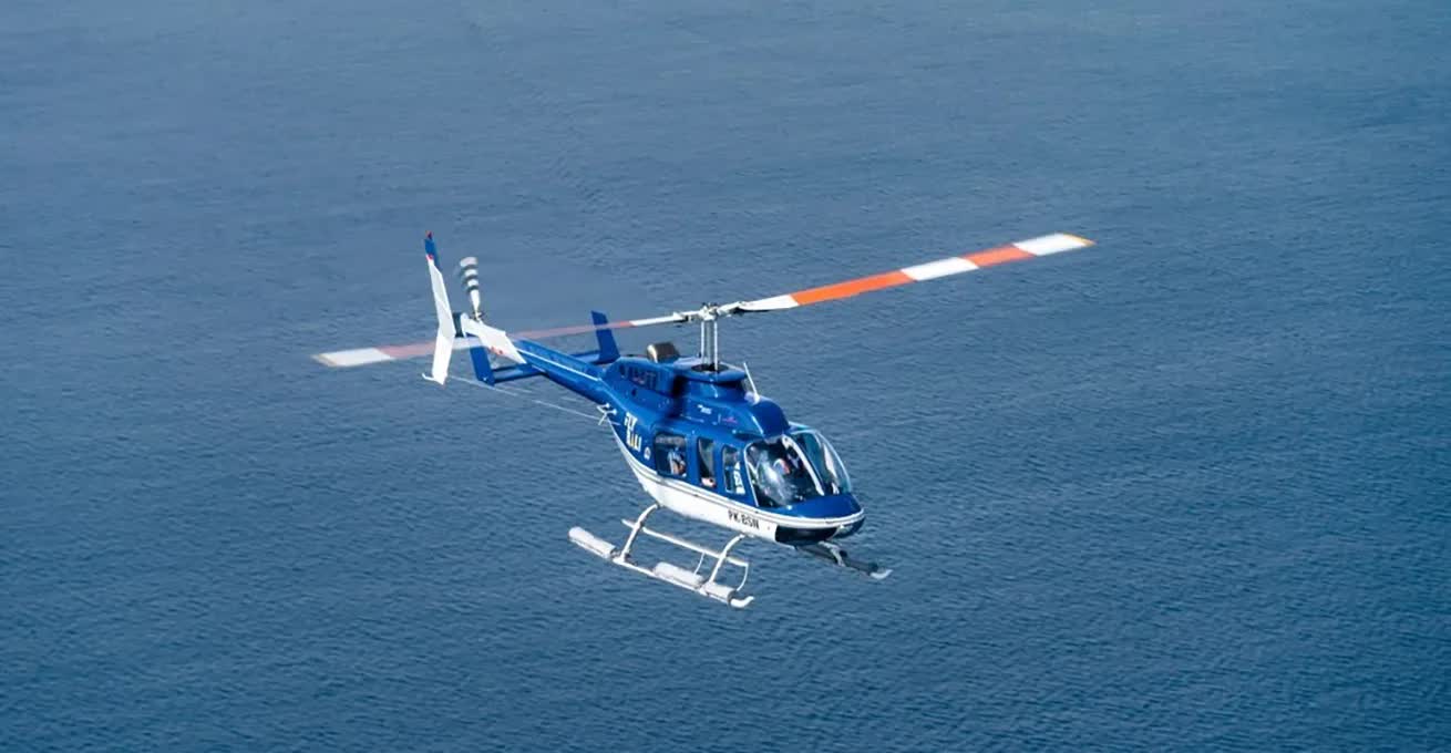 A blue and white helicopter from Fly Bali Heliport company is flying over the water